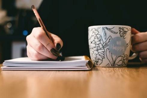 Image displays a person holding a mug and writing in a notebook.