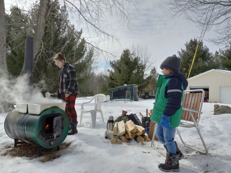 A snowy outdoor scene with a large barrel that is open on one end with a fire inside and a chimney on one end. On top sit two steaming pans. An adult is standing near the barrel with a spoon. Nearby, a pile of wood and a smiling child dressed in snow gear.