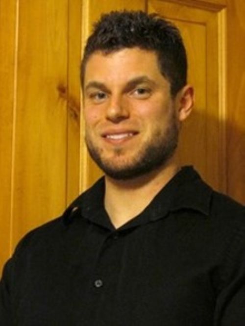 A headshot of Tyler Becker smiling, wearing a black shirt in front of a wood-paneled wall