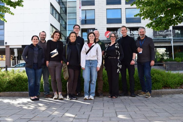 A group of people posing for a picture. There are multi-story buildings in the background with green shrubs and trees in the background.