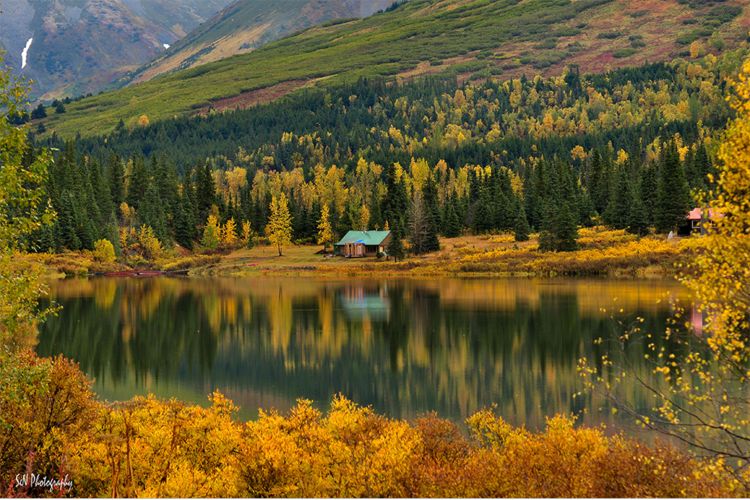 A log cabin in the distance sitting behind a lake with foliage and hills behind.