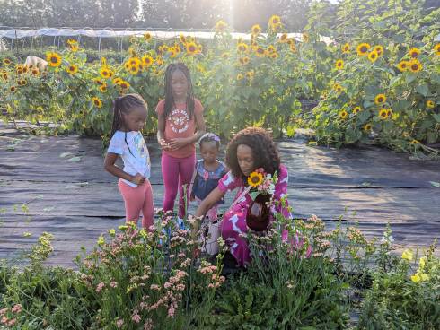 A woman and three girls pick a bouquet of flowers in a field with sunflowers in the background
