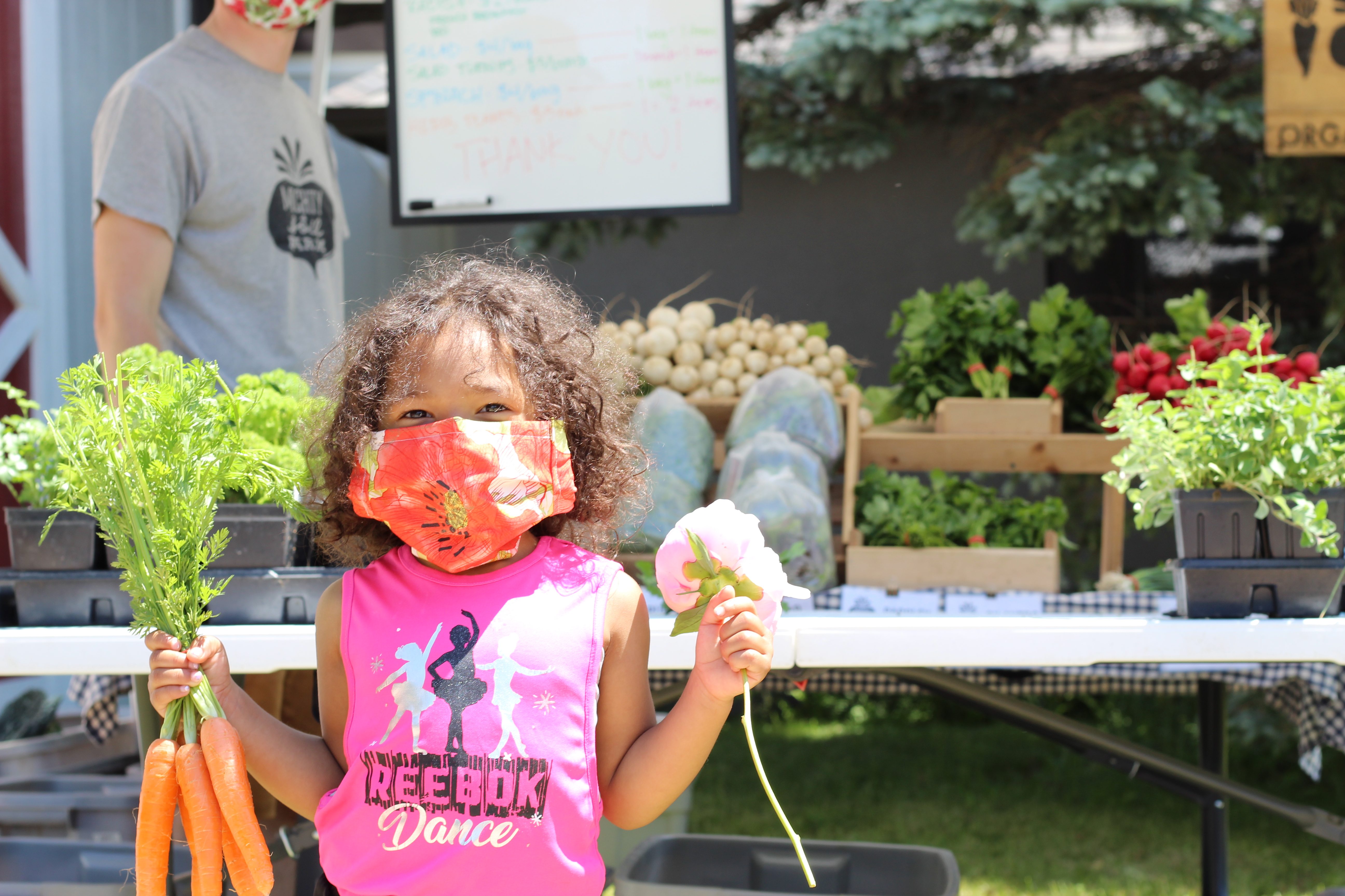 Young girl wearing a mask holds a flower and a bunch of carrots in front of a farmers market stall.